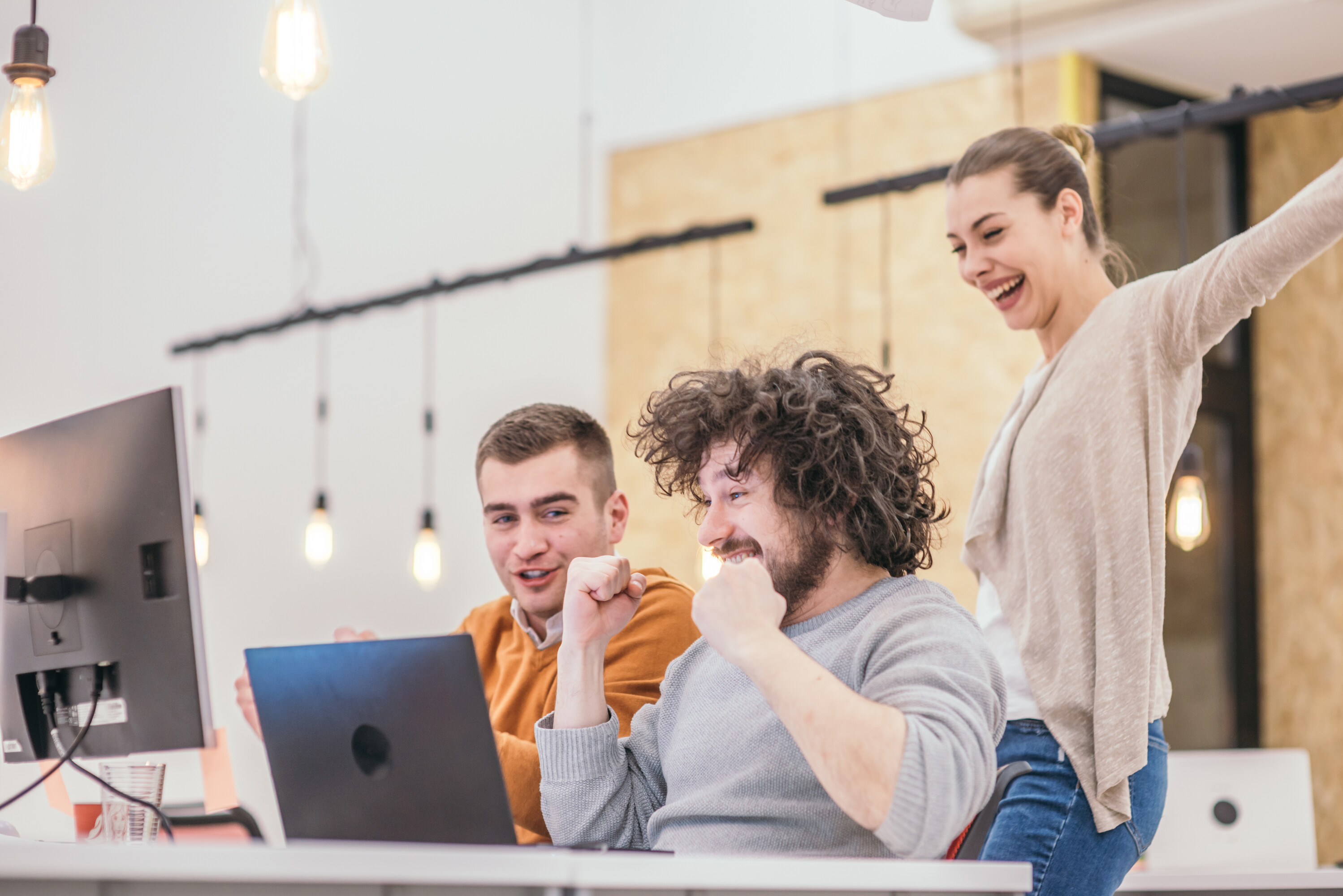 Happy surprise for the teammates. Coworkers cheering while looking at a computer, screen at the office. Positive vibe at the startup modern office.