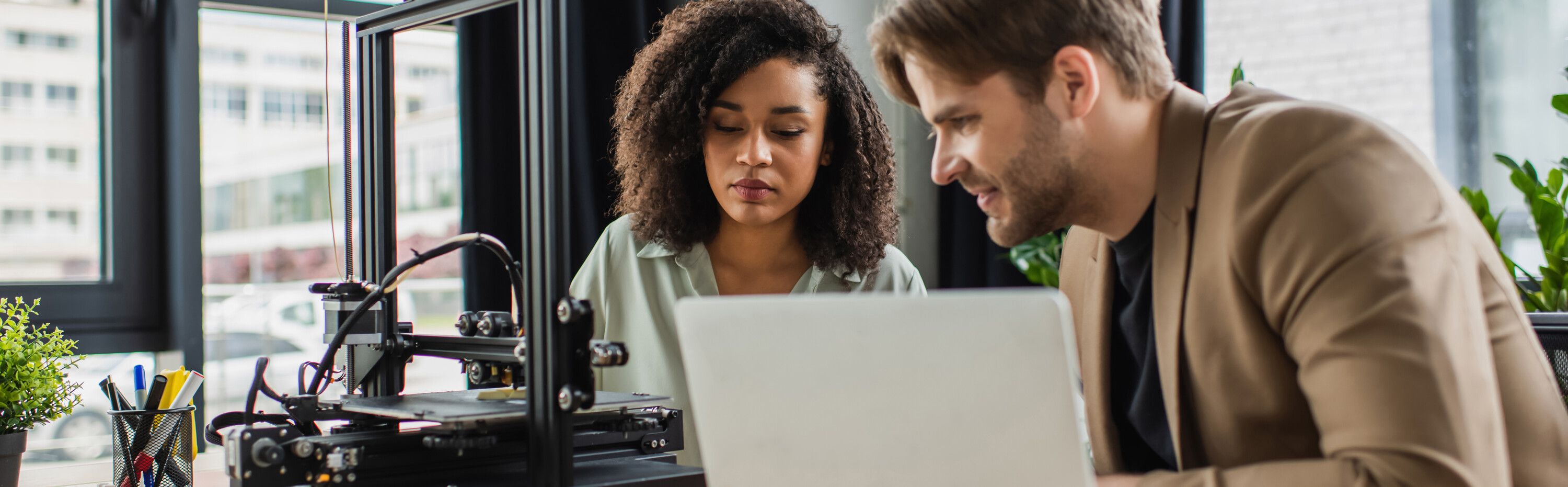 smiling young man sitting with african american colleague near 3D printer and laptop in modern office, banner